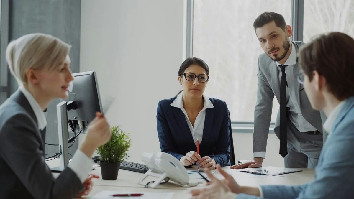Four professionals in business attire in a serious discussion around a meeting table, papers and a monitor in front of them.