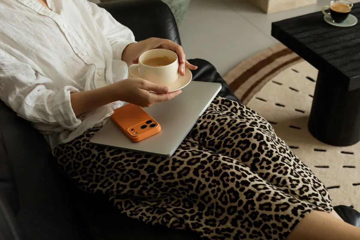 A woman on a sofa holding a coffee cup, with a closed laptop and an orange phone on her lap.