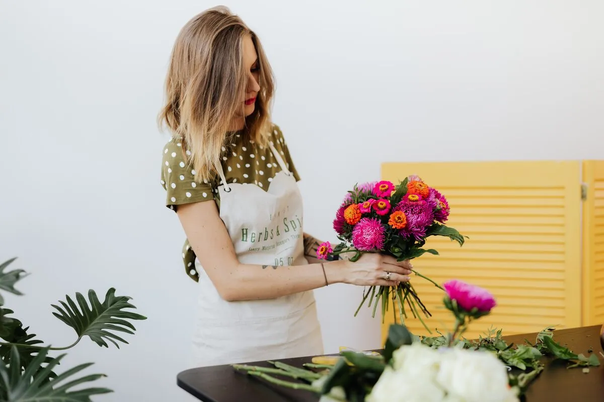 A florist in an apron arranging a bright bouquet of zinnias and asters at her bench.