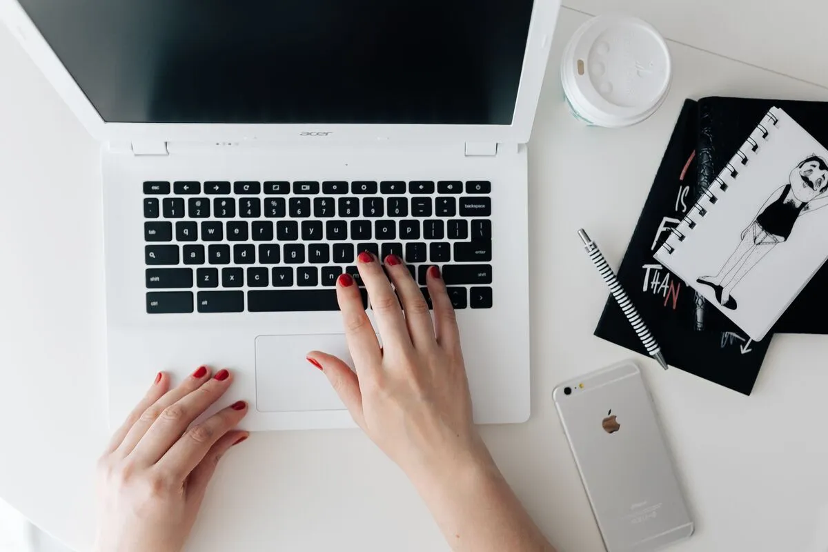 Overhead shot of a woman's hands typing on a white laptop beside a coffee cup, notebook and phone.
