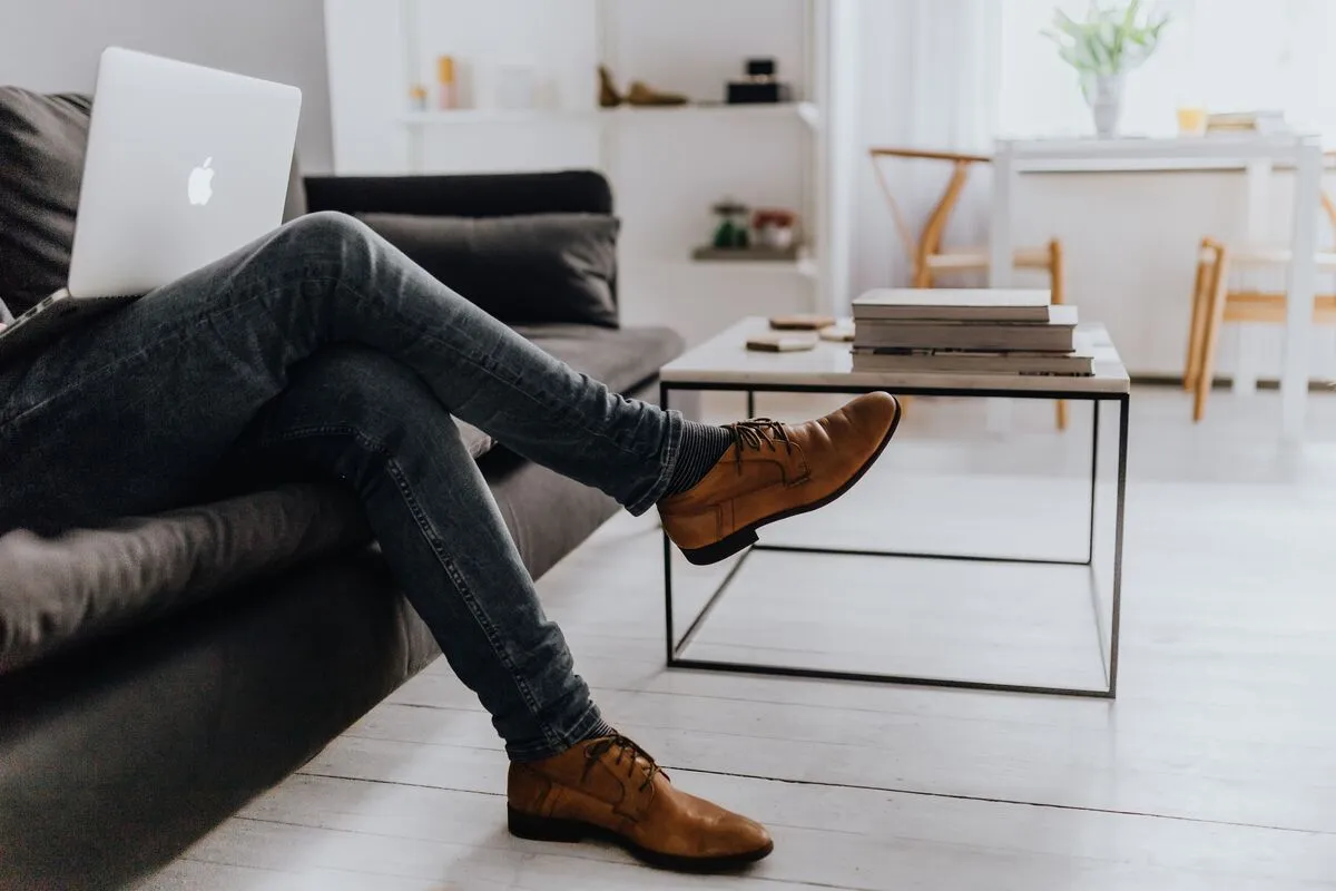 A man's legs stretched out on a sofa with a laptop balanced on his lap in a bright Scandinavian apartment.