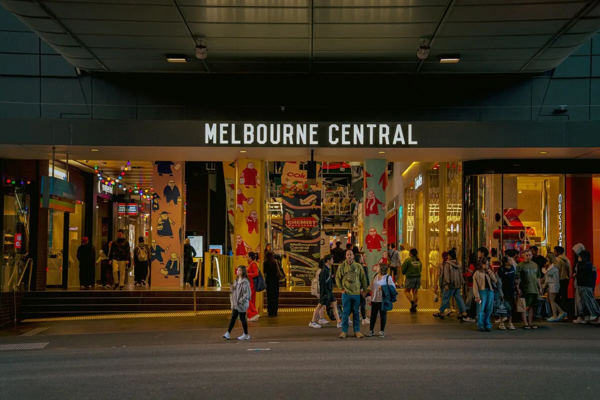 Crowds pass colourful lit shopfronts at the Melbourne Central entrance at night.