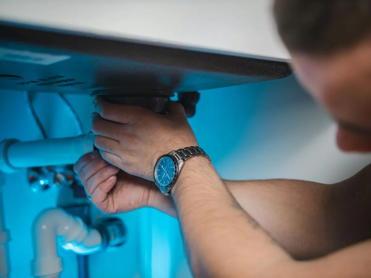A plumber's hands working on the waste trap under a sink, lit by a blue work light.