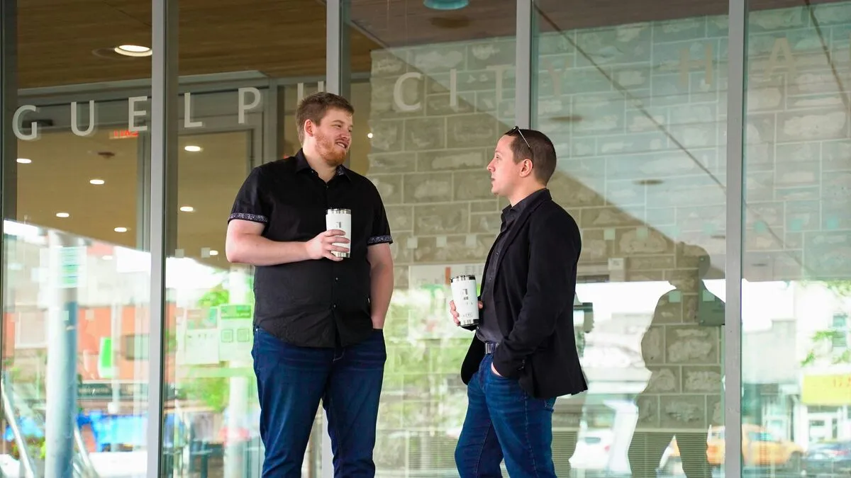 Two men in business-casual chatting with takeaway coffees outside a modern glass office.
