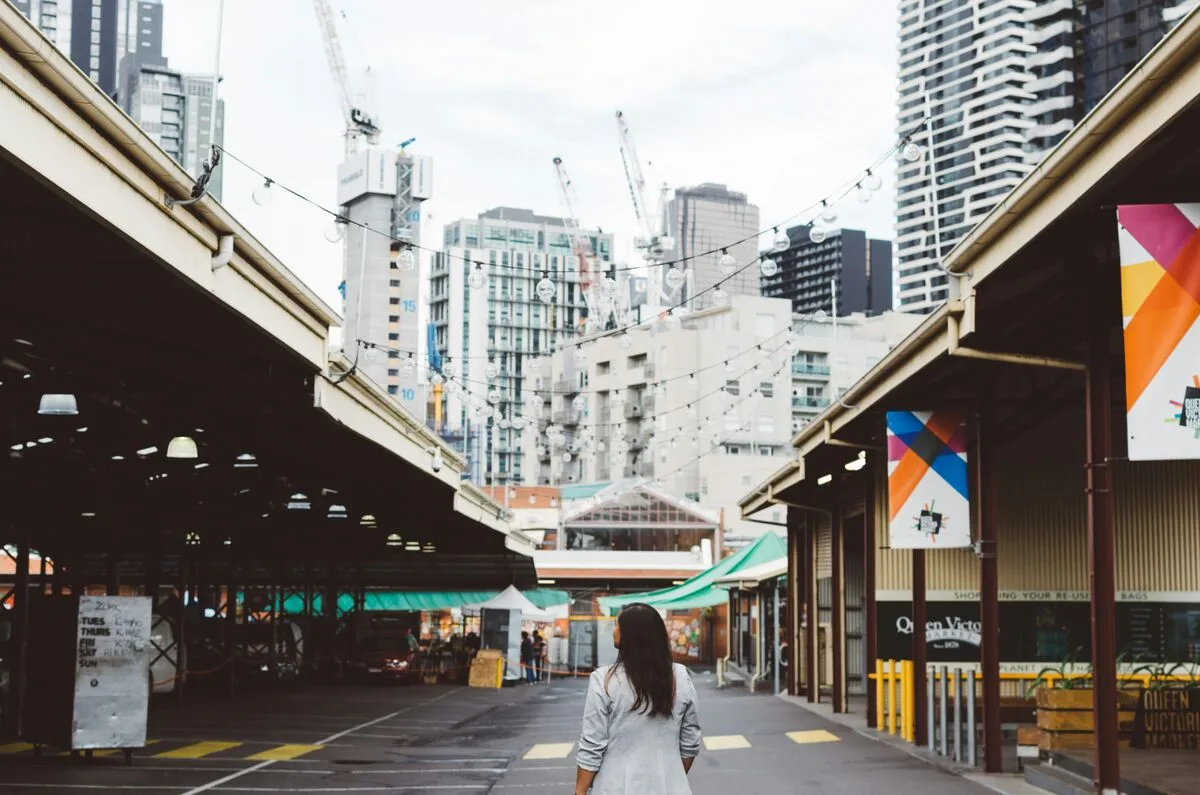 A person walks through Melbourne's Queen Victoria Market past shopfronts and string lights, with city cranes on the skyline behind.