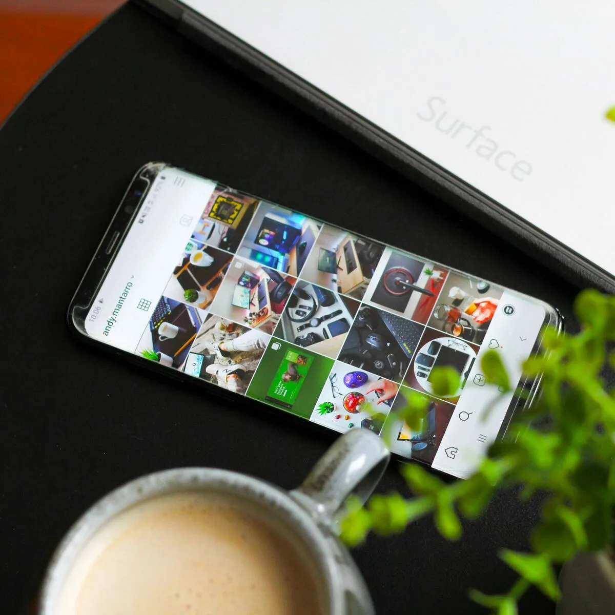 A smartphone on a Surface laptop showing an Instagram photo grid, beside a coffee and a plant.