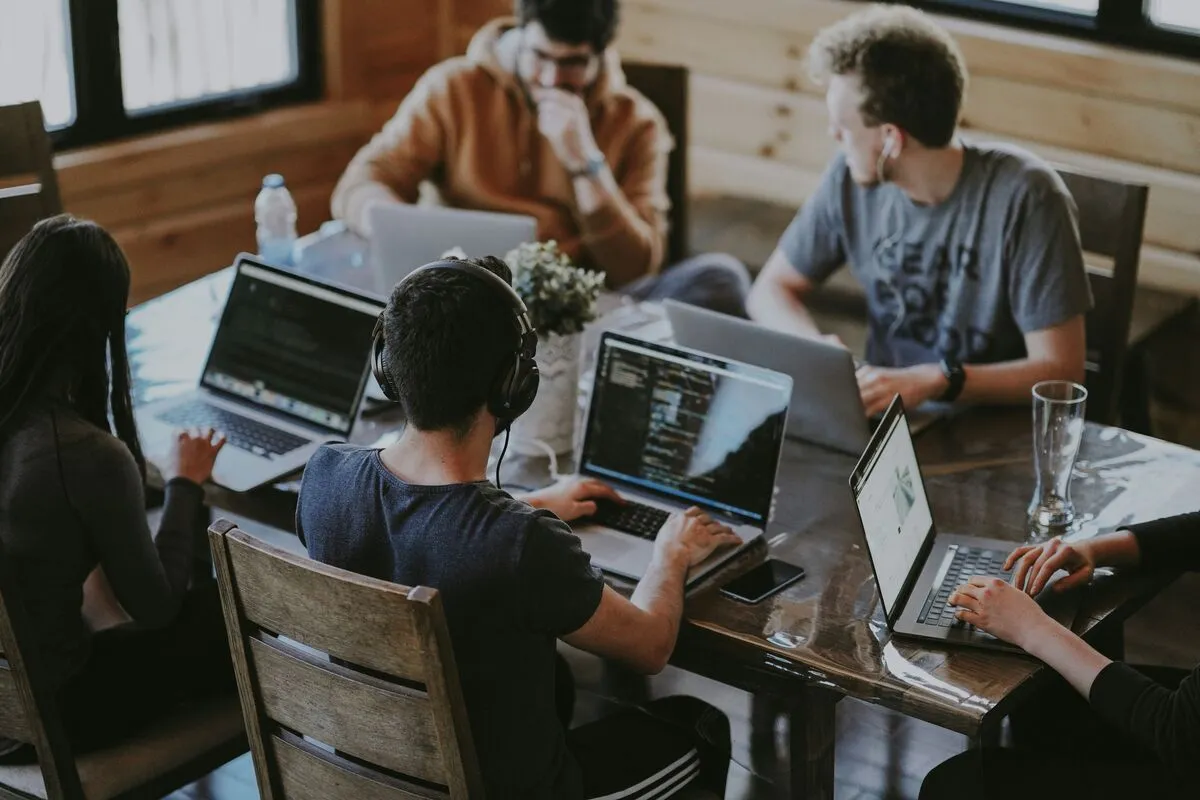 A team of people working head-down on laptops around a wooden table, one wearing headphones.