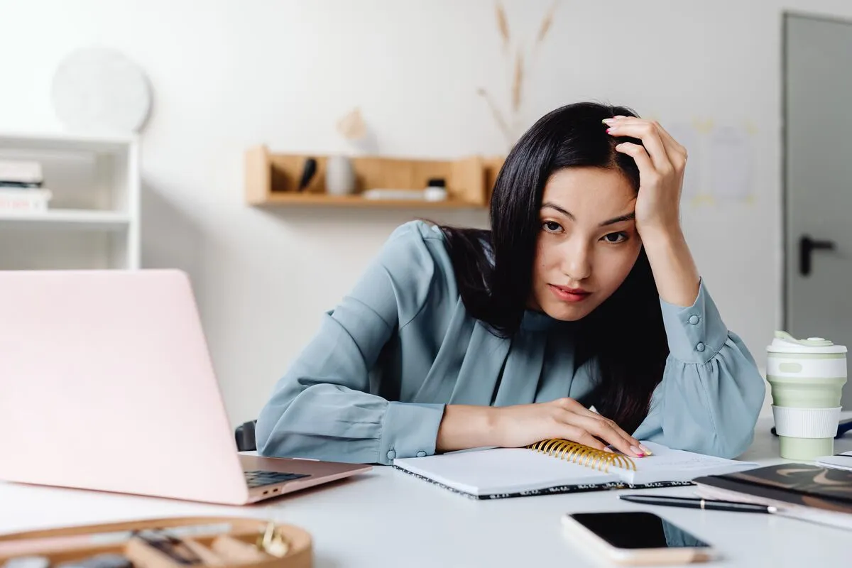 A woman at a white desk resting her head on her hand, looking fed up beside a pink laptop and notebook.