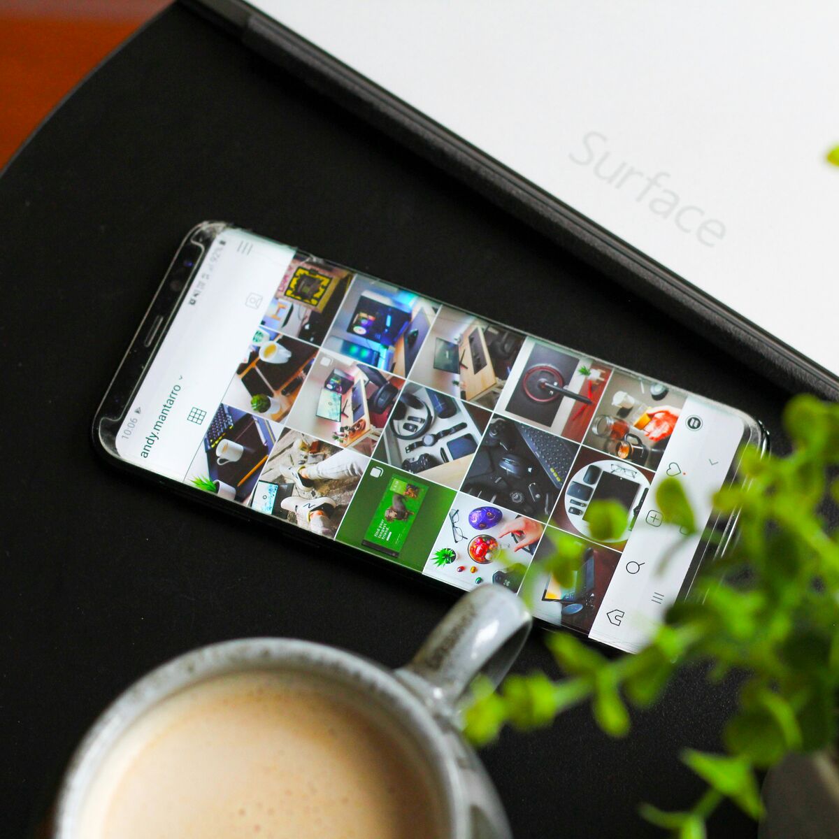 A smartphone on a Surface laptop showing an Instagram photo grid, beside a coffee and a plant.